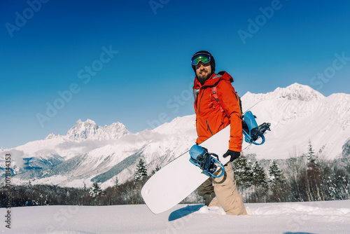 snowboarder holding snowboard walking through powder snow against backdrop of sunny mountain landscape on winter day
