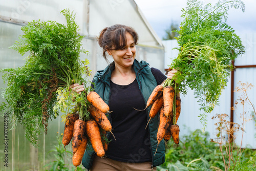 Happy young female farmer harvesting carrots in vegetable garden in autumn time