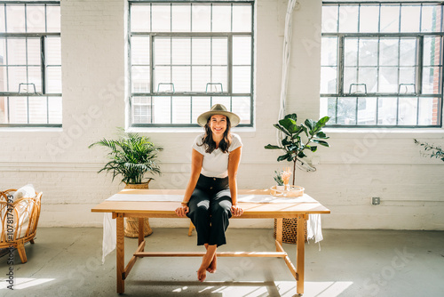 Smiling woman in hat sitting on table in bright, plant-filled room photo