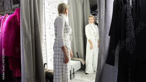 Stylish Woman Trying On and Selecting Fashionable Clothing in Fitting Room of Female Fashion Store, Featuring Chic Checkered White Suit While Looking in Mirror