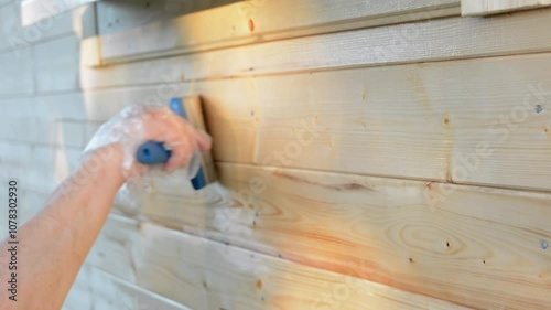 A hand wearing a protective plastic glove applies wood stain with a paintbrush to a wooden wall, focusing on even coverage and weather protection outdoors