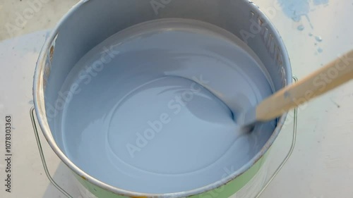 A close-up of gray paint being stirred smoothly in a paint can with a wooden stir stick. The paint swirls evenly as it is prepared for application in a DIY or home improvement project.