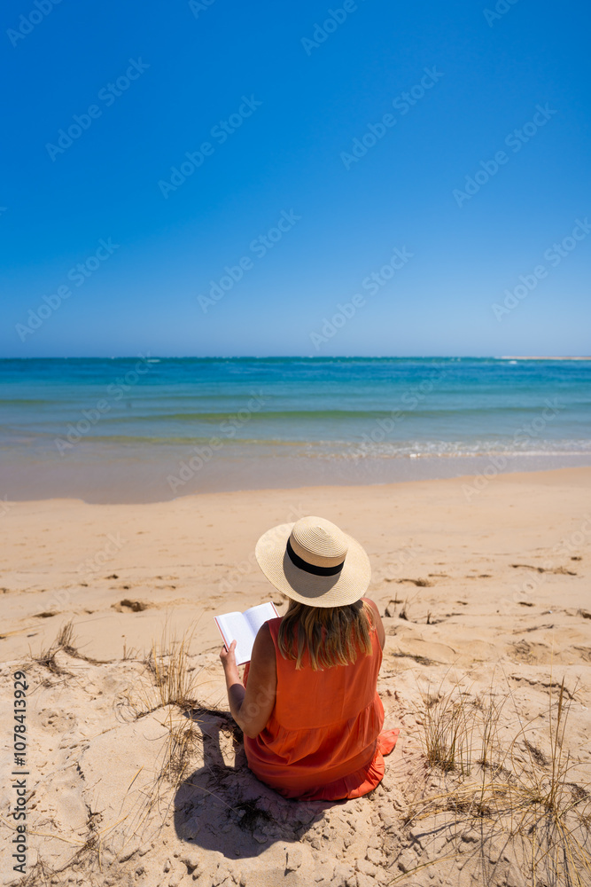 Beautiful middle aged woman wearing orange dress, sunglasses and sun hat sitting on sandy beach and reading book on sunny summer day. Back view. Tourist on best beach. Beautiful middle aged woman wearing orange dress, sunglasses and sun hat sitting on sandy beach and reading book on sunny summer day. Back view. Tourist on best beach.