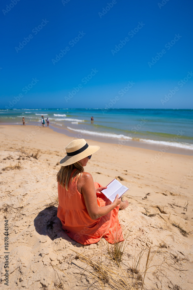 Φωτογραφία Beautiful middle aged woman wearing orange dress, sunglasses and sun hat sitting on sandy beach and reading book on sunny summer day