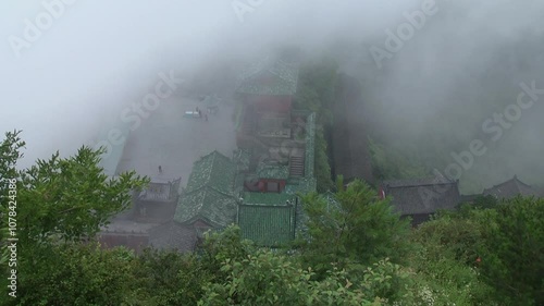 Misty view of a Taoist temple complex nestled in the Wudang Mountains, China. Ancient green-tiled rooftops and courtyards are partially obscured by clouds, creating a mystical, serene atmosphere.