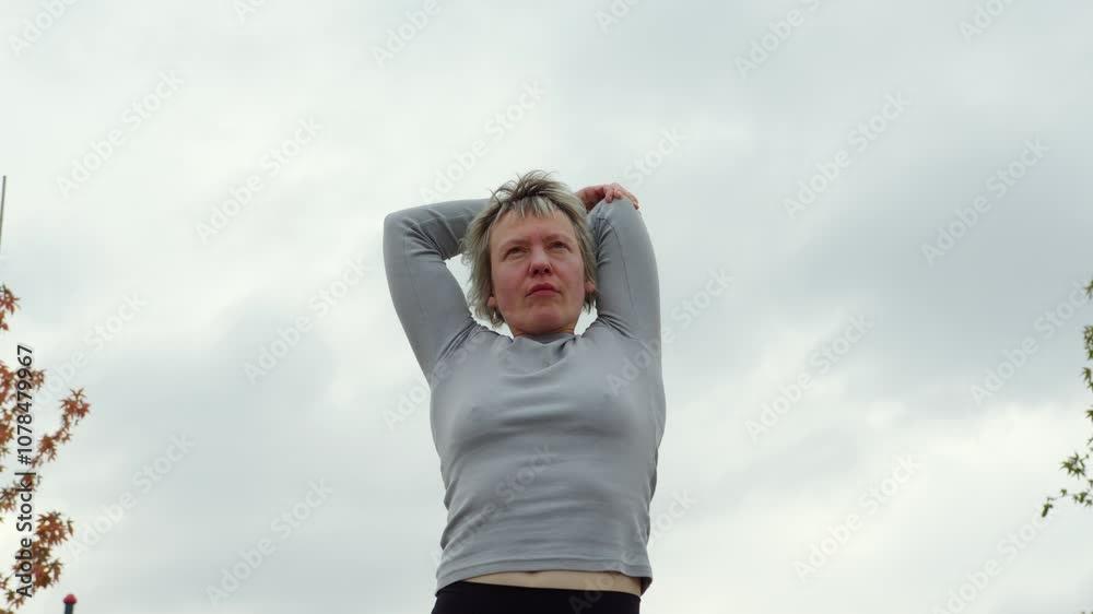 A woman with short blonde hair stretching her arms outdoors under a cloudy sky. She appears focused and calm, wearing a gray top, with autumn leaves partially visible on the side. Fitness and wellness A woman with short blonde hair stretching her arms outdoors under a cloudy sky. She appears focused and calm, wearing a gray top, with autumn leaves partially visible on the side. Fitness and wellness