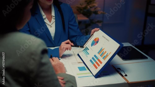 Businesswoman in a blue blazer points at charts on a clipboard while explaining a strategy to her business partner at the office