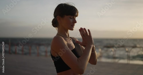 Close up of a happy brunette girl in a black sports summer uniform folds her hands in front of her chest and begins her yoga and stretching session by moving her foot forward and meditating near the