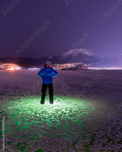 Person Standing On A Frozen Lake Dillon In Colorado At Night With The Ice Illuminated Below And The Mountain Town Of Wilderness, Colorado In The Distance photo