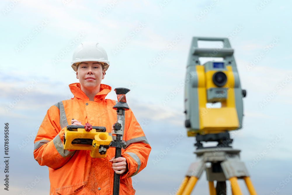 Woamn surveyor in orange jacket using equipment at construction site during cloudy day Woamn surveyor in orange jacket using equipment at construction site during cloudy day