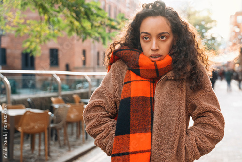 Upset young woman wearing a cozy tan jacket and orange plaid scarf stands in a vibrant urban street during autumn