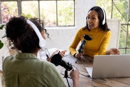 Two multiracial female friends recording podcast at home, engaging in lively conversation photo