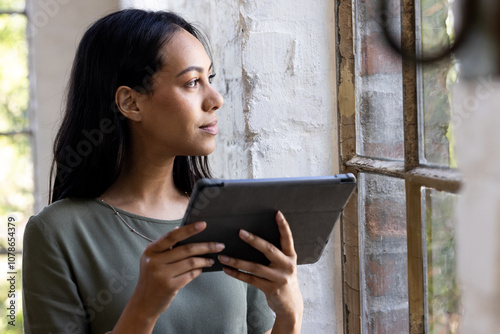 Holding tablet, woman gazing thoughtfully out window in home setting