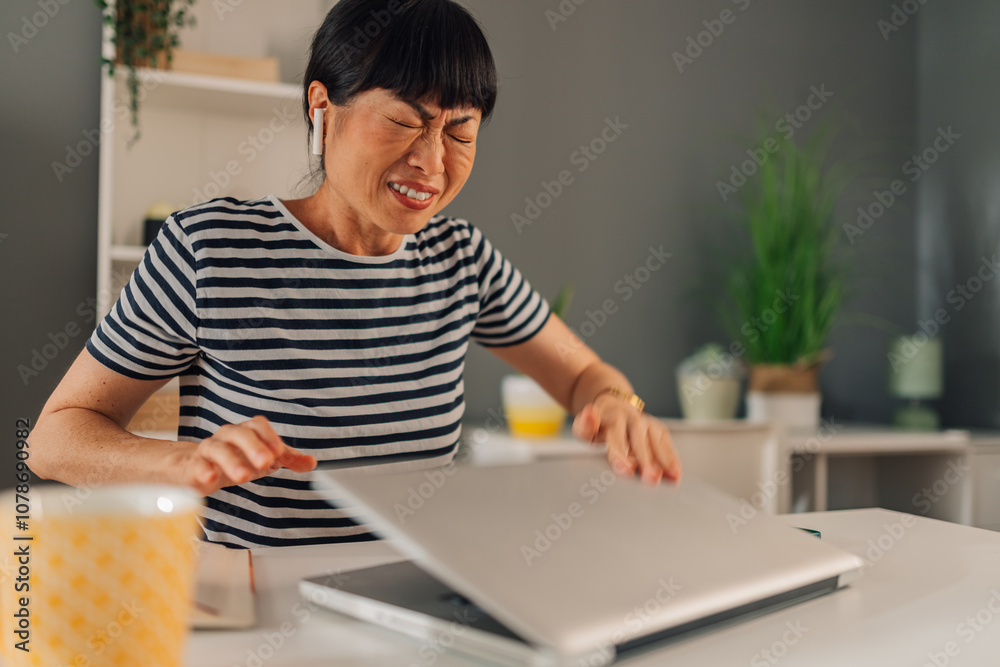 Angry asian woman closing a laptop on desk with striped shirt, mug and book. Angry asian woman closing a laptop on desk with striped shirt, mug and book.