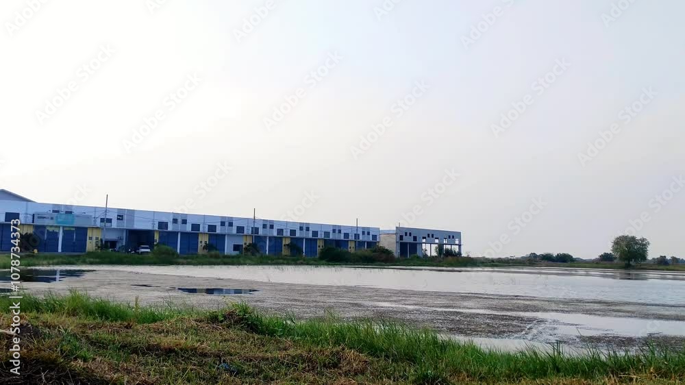 A warehouse complex stands near a calm pond, with grassy edges framing the water. The sky is clear, and the still pond surface lacks any reflections. A warehouse complex stands near a calm pond, with grassy edges framing the water. The sky is clear, and the still pond surface lacks any reflections.