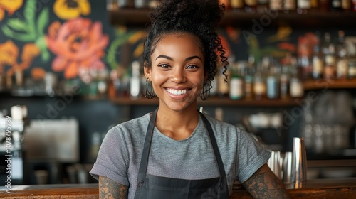 A woman with a smile on her face stands behind a bar. She is wearing a black apron and a gray shirt