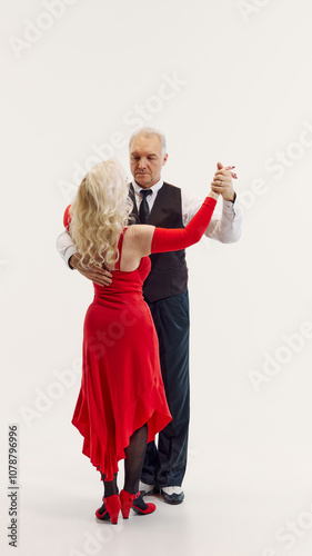 Elegant senior blonde woman in red dress and handsome senior man in black suit dancing tango isolated on white background. Concept of retro dance, hobby, aging, active lifestyle photo