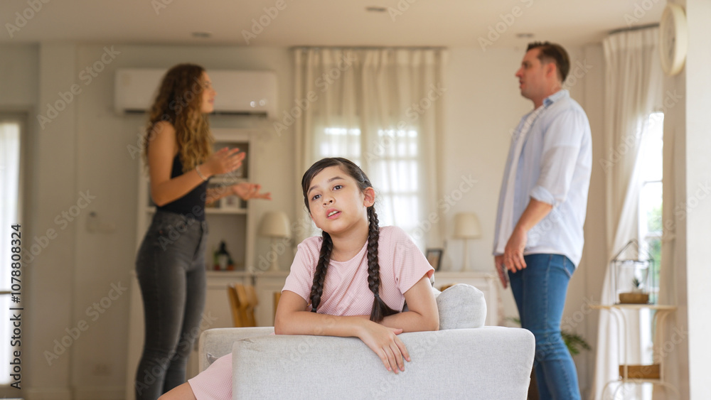 Annoyed and unhappy young girl sitting on sofa trapped in middle of tension by her parent argument in living room. Unhealthy domestic lifestyle and traumatic childhood develop to depression Synchronos Annoyed and unhappy young girl sitting on sofa trapped in middle of tension by her parent argument in living room. Unhealthy domestic lifestyle and traumatic childhood develop to depression Synchronos