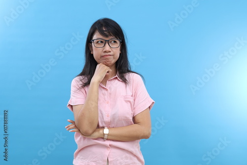 Grumpy displeased Asian female smirks face, keeps hands half crossed, holds chin, has angry expression, being deep in thoughts, dressed in casual wear, models over blue wall, copy space aside photo