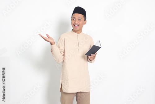 Portrait of excited muslim man in koko shirt with peci reading a notebook. Happy Asian guy pointing to the side with finger. Isolated image on white background photo