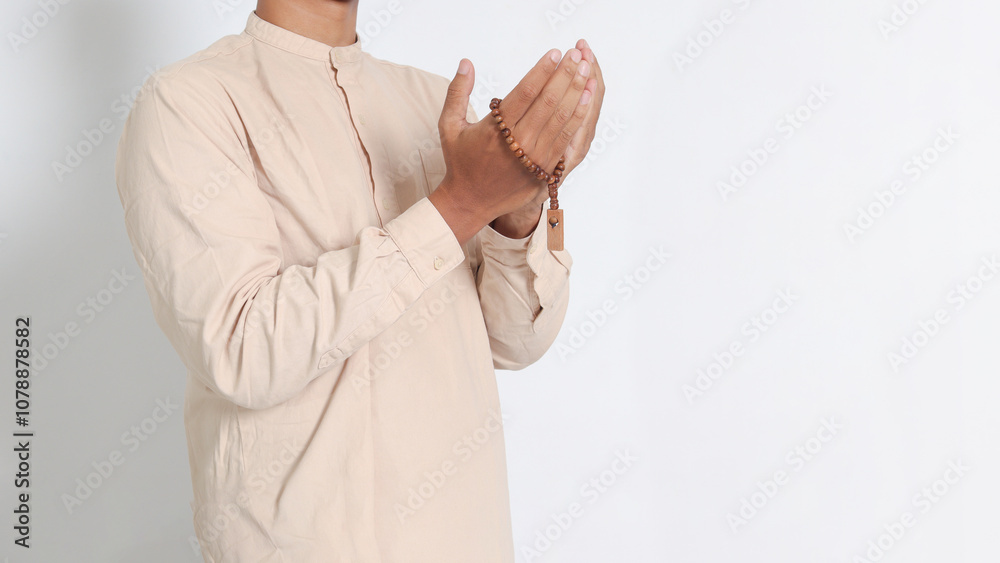 Close up portrait of religious Asian muslim man in koko shirt with skullcap praying earnestly with his hands raised, holding islamic beads. Devout faith concept. Isolated image on white background