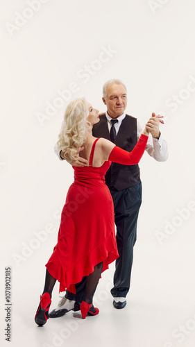 Elegant senior blonde woman in red dress and handsome senior man in black suit dancing tango isolated on white background. Concept of retro dance, hobby, aging, active lifestyle photo