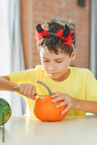 Boy making jack o lantern at home white kitchen, cutting real pumpkins and playing with paper black ghosts, lovely vibe of halloween