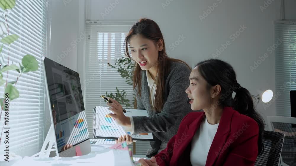Two asian businesswomen stand in an office, discussing charts and data displayed on a desktop computer Two asian businesswomen stand in an office, discussing charts and data displayed on a desktop computer