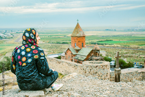 Young Woman on viewpoint wear modest outfit sit enjoy travel visit sightseeing famous Khor Virap monastery building and chapel. Travel and religious destinations in Armenia concept photo