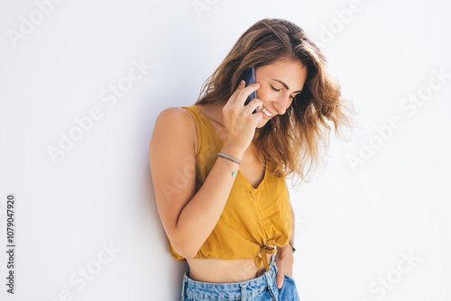 Smiling modern fit young woman in casual outfit talking on mobile while leaning on white wall photo