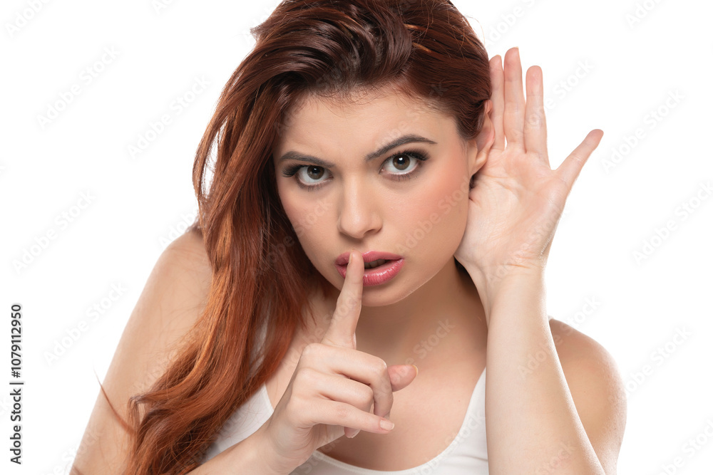 Redhead woman holds her hand near ear and listens carefully with silence gesture isolated on white background