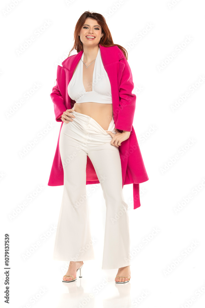 Fashion style studio portrait of beautiful young woman. White wide leg pants and pink coat. Model sitting and standing against white background Fashion style studio portrait of beautiful young woman. White wide leg pants and pink coat. Model sitting and standing against white background