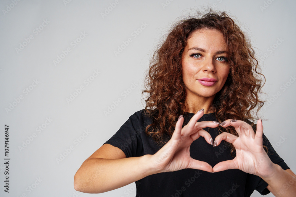 A young woman with curly hair makes a heart shape with her hands against a neutral backdrop A young woman with curly hair makes a heart shape with her hands against a neutral backdrop