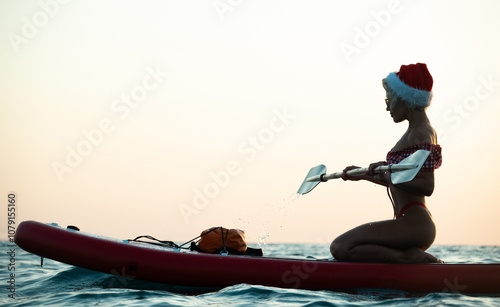 Sexy girl in a Santa Claus hat floats on the sea on a Sup Board in a red bikini celebrating Christmas at a resort in a hot country photo