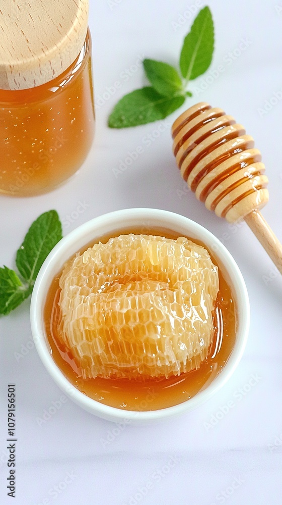 A white surface with green leaves holds a bowl of honey beside a jar of honey and a wooden honeycomb A white surface with green leaves holds a bowl of honey beside a jar of honey and a wooden honeycomb
