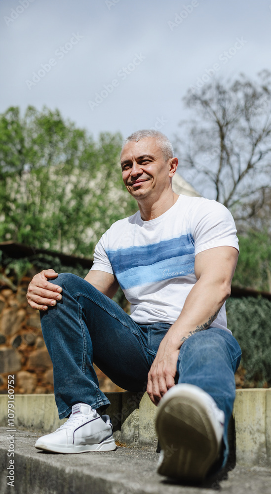 A man is sitting on a cement ledge wearing a white shirt and blue jeans. He is smiling and he is enjoying the moment A man is sitting on a cement ledge wearing a white shirt and blue jeans. He is smiling and he is enjoying the moment