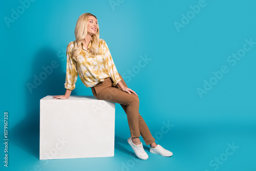 Full length photo of attractive mature woman sit cube look empty space dressed stylish formalwear clothes isolated on cyan color background photo