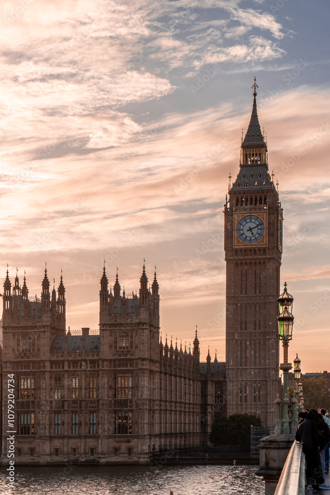 Fototapeta premium Scenic view of Big Ben and the Palace of Westminster along the Thames River, beautifully illuminated by a warm sunset sky.