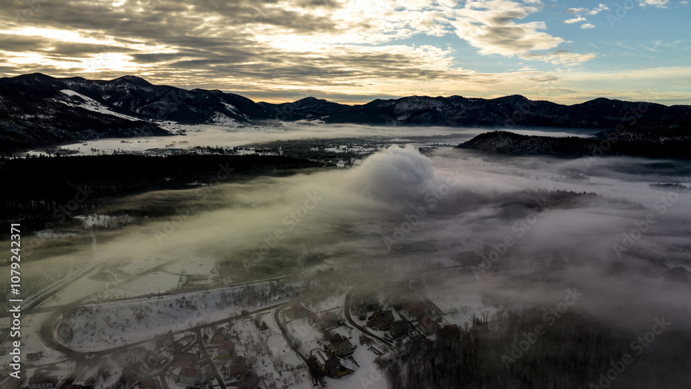 Aerial view of fog covering a snowy land with terrain in the background under cloudy sky at dawn Aerial view of fog covering a snowy land with terrain in the background under cloudy sky at dawn