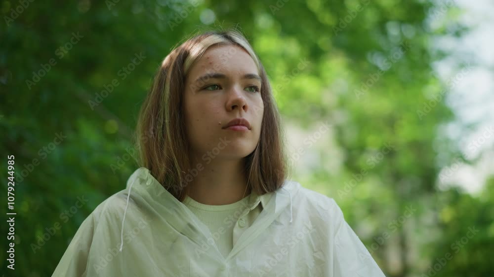 Young woman in translucent raincoat stands outdoors, lost in deep thought, with a serene and reflective expression on her face, surrounded by lush green trees, she appears contemplative Young woman in translucent raincoat stands outdoors, lost in deep thought, with a serene and reflective expression on her face, surrounded by lush green trees, she appears contemplative