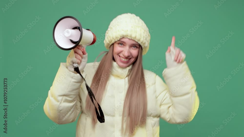 A blonde woman in warm yellow jacket and hat on green background holds a loudspeaker in her hands and shouts