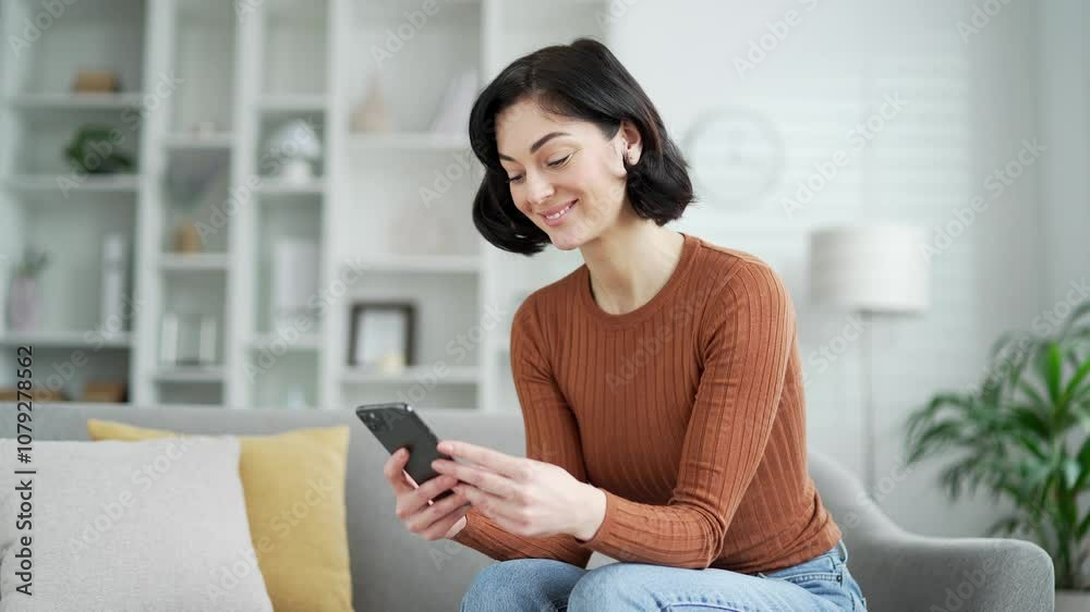 Young adult woman in cozy living room sitting on sofa, smiling at phone, enjoying casual time at home. Smiling female student browses social networks, makes purchases, chats, writes or reads messages Young adult woman in cozy living room sitting on sofa, smiling at phone, enjoying casual time at home. Smiling female student browses social networks, makes purchases, chats, writes or reads messages