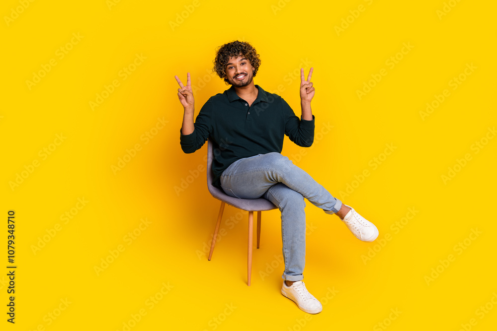 Young man sitting on chair against vibrant yellow background making peace signs with joyful expression Young man sitting on chair against vibrant yellow background making peace signs with joyful expression