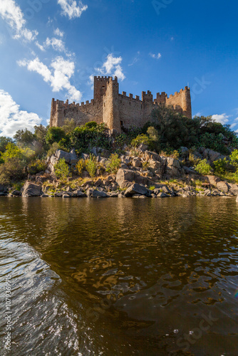 A historic castle stands majestically atop a rocky island surrounded by calm river waters, with a backdrop of lush hills