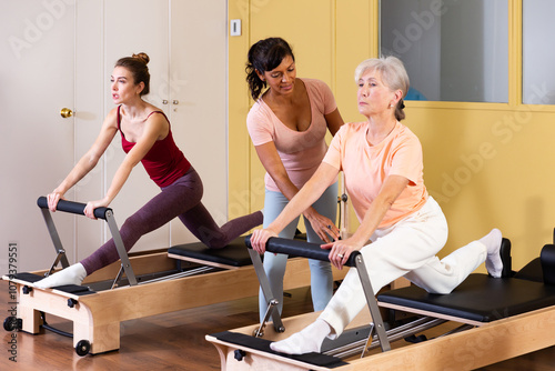 Two active women practicing Pilates in a fitness studio perform an exercise using a reformer bed, where a female instructor ..helps them do it correctly photo