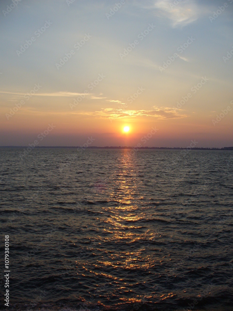 Blue sky with orange and yellow sunset over horizon of dark water surface with reflection of sunlight on water with small waves - natural evening scene. Blue sky with orange and yellow sunset over horizon of dark water surface with reflection of sunlight on water with small waves - natural evening scene.