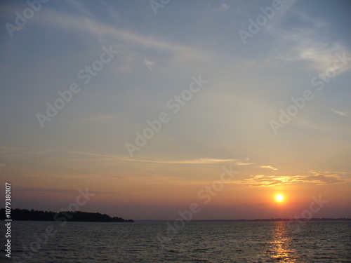 Blue sky with orange and yellow sunset over horizon of dark water surface with reflection of sunlight on water with small waves - natural evening scene. Topics: weather, natural environment, season