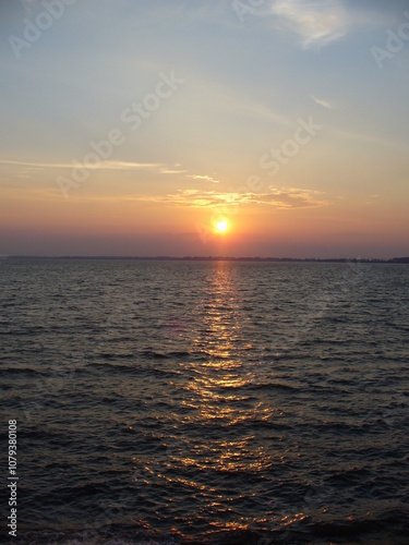 Blue sky with orange and yellow sunset over horizon of dark water surface with reflection of sunlight on water with small waves - natural evening scene.