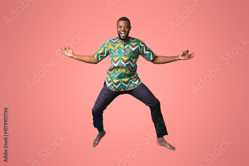 Carefree black man in african costume posing while jumping up on blue studio background. Smiling african american guy wearing traditional clothes, full size photo. Traditions and culture concept