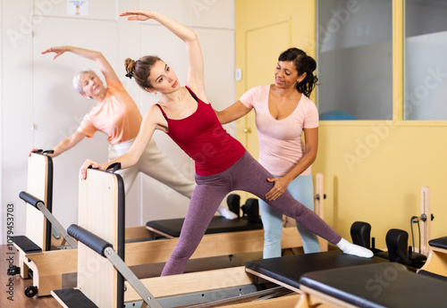 Friendly woman Pilates instructor, conducting a workout in a fitness studio, helps the female group to perform a gymnastic ..stretching exercise photo